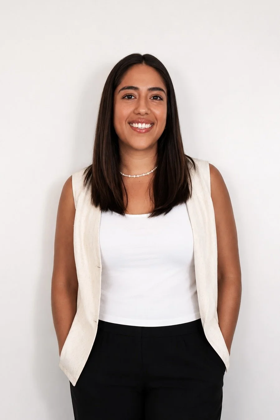 A young woman with straight dark hair, smiling, wearing a white sleeveless top, a beige vest, black pants, a pearl necklace, standing against a plain white background.