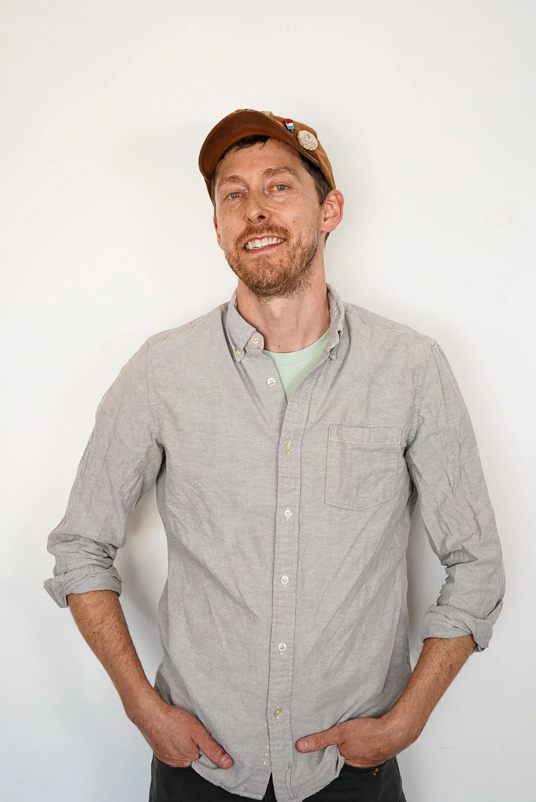 A man with a beard and short hair, wearing a brown cap with pins, a light gray button-up shirt, and dark pants, standing in front of a plain white background.