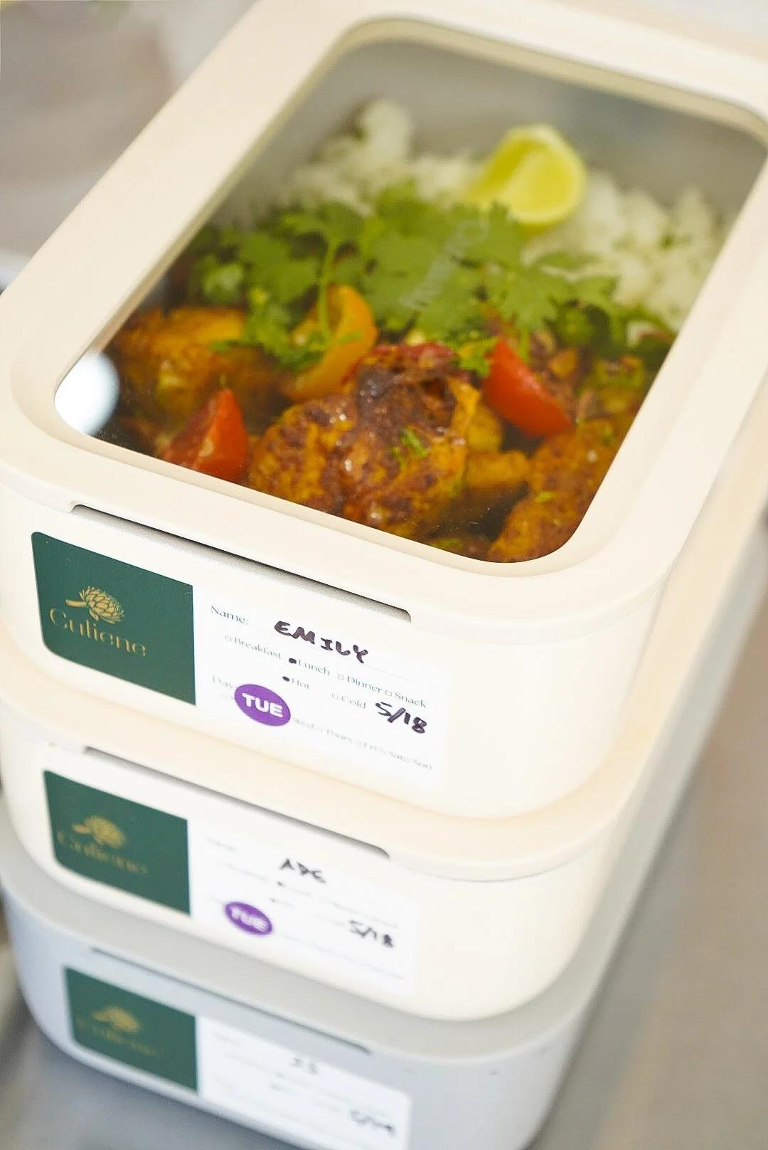 A food container with a clear lid showing a meal of meatballs with sauce, cherry tomatoes, rice, lemon, and cilantro.