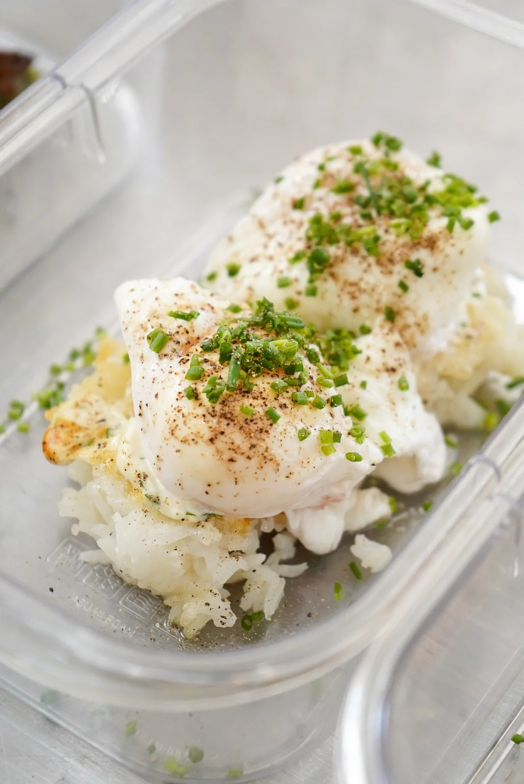 Poached eggs topped with chopped green onions and black pepper, served on a bed of mashed potatoes in a clear food storage container.