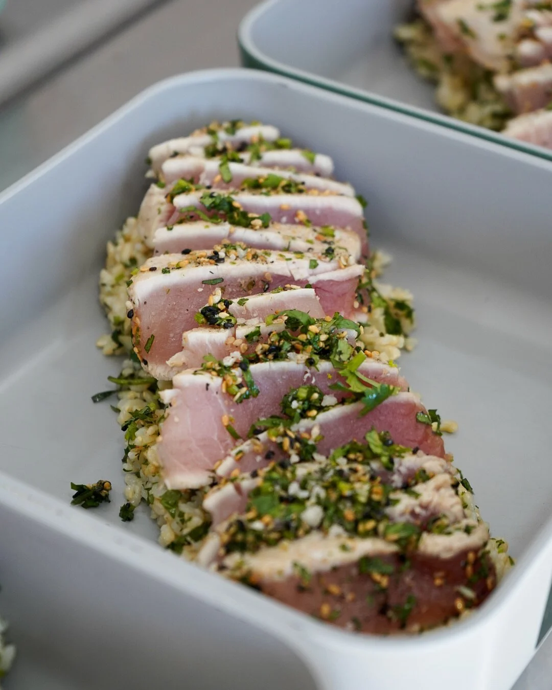 Sliced raw tuna tuna with green herbs and crushed nuts on a bed of rice in a white container.