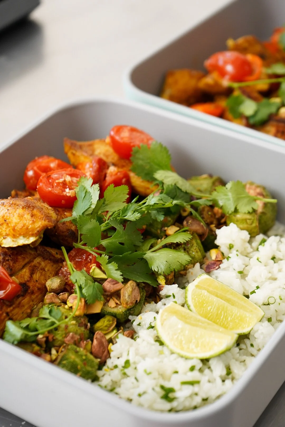 Close-up of a white food container filled with rice, grilled chicken, cherry tomatoes, jalapeño slices, chopped pistachios, fresh cilantro, and lime wedges.