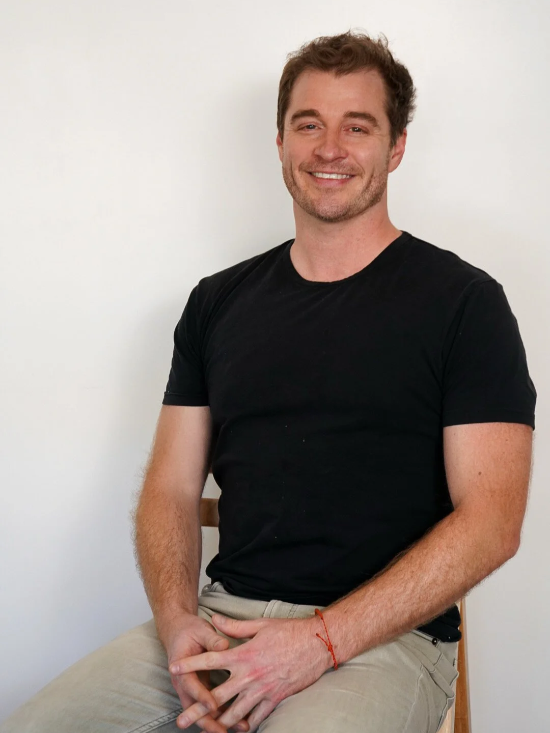 A smiling man with brown hair, wearing a black t-shirt and beige pants, sitting on a wooden chair against a plain white wall.