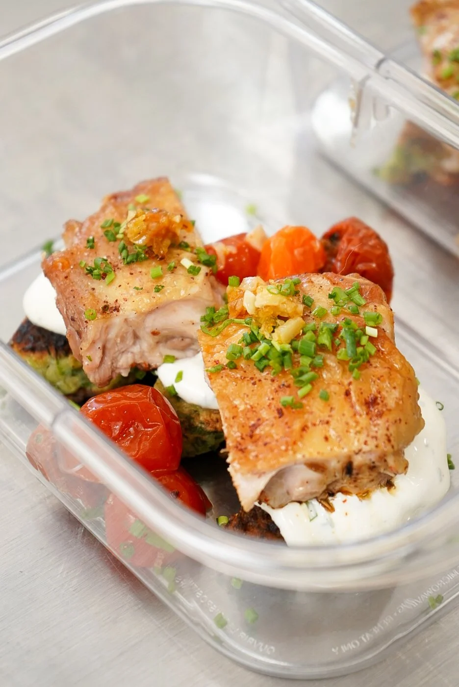 Cooked fish fillets topped with chopped green onions and garlic, served over vegetables with cherry tomatoes, garnished with herbs and a creamy sauce in a glass container.