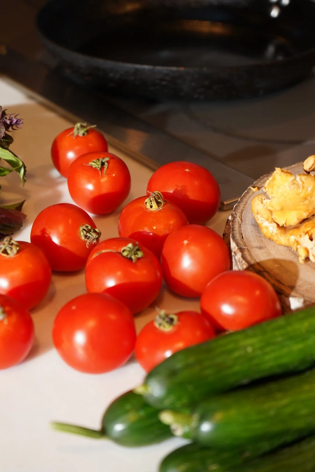 Group of fresh red tomatoes on a kitchen counter next to cucumbers and a wooden bowl with ginger.