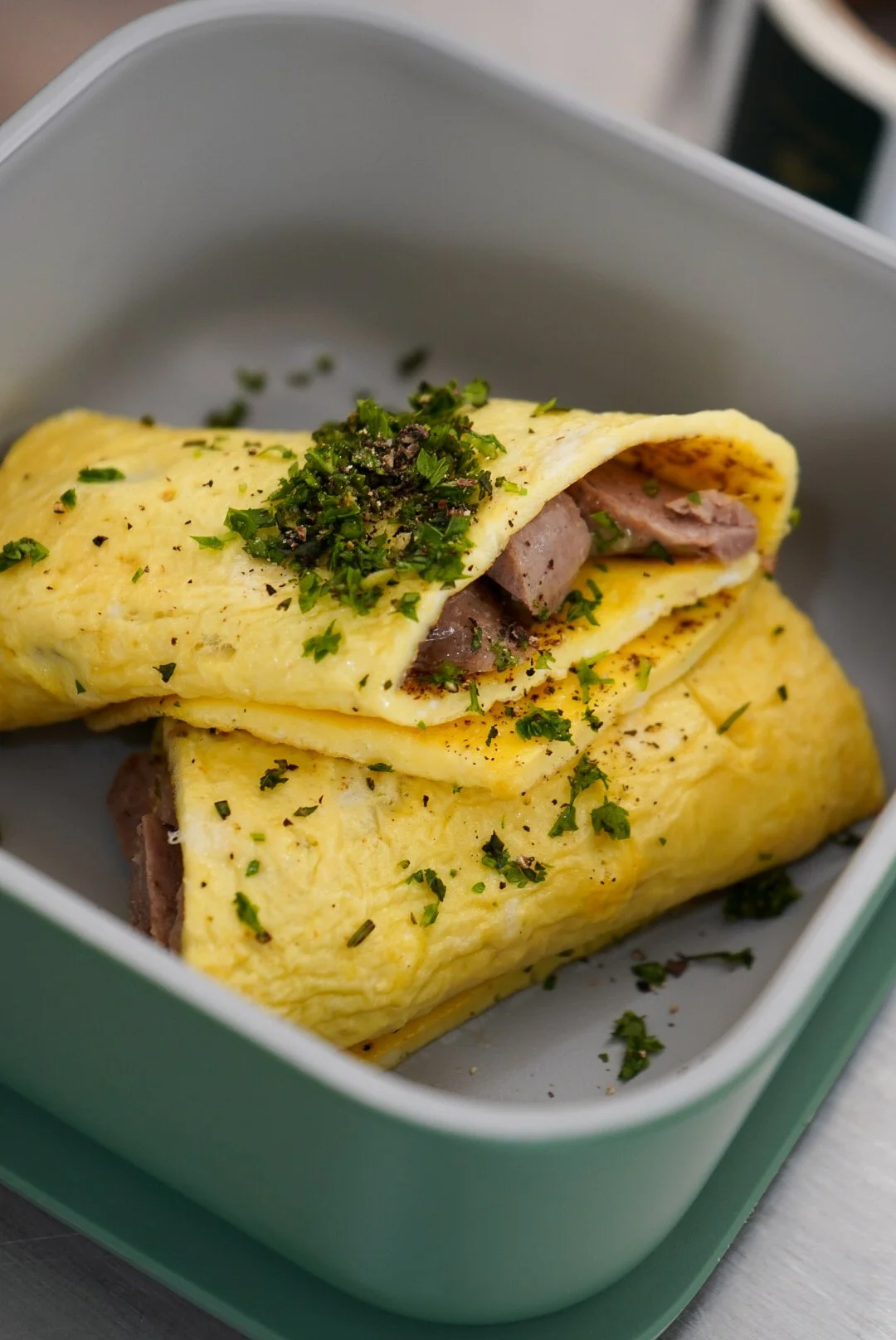A folded yellow omelette stuffed with pieces of cooked ham, topped with chopped herbs and black pepper, served in a white container.