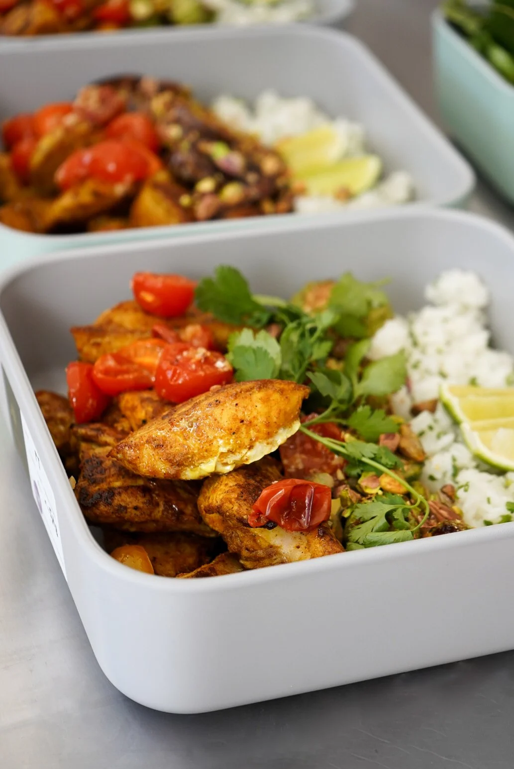 Meal prep containers with seasoned grilled chicken, cherry tomatoes, rice, and fresh cilantro with lemon wedges.