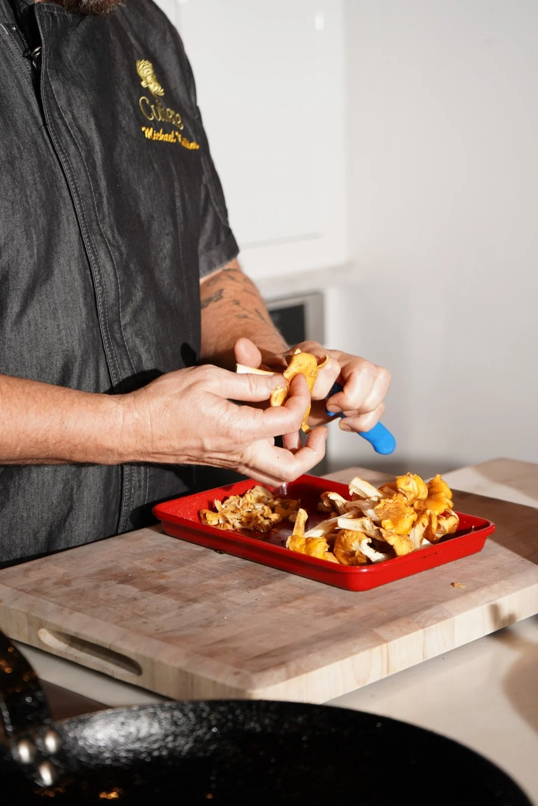 Person with tattooed arms peeling the outer layer of a golden-colored mushroom over a red tray filled with more mushrooms on a wooden cutting board.