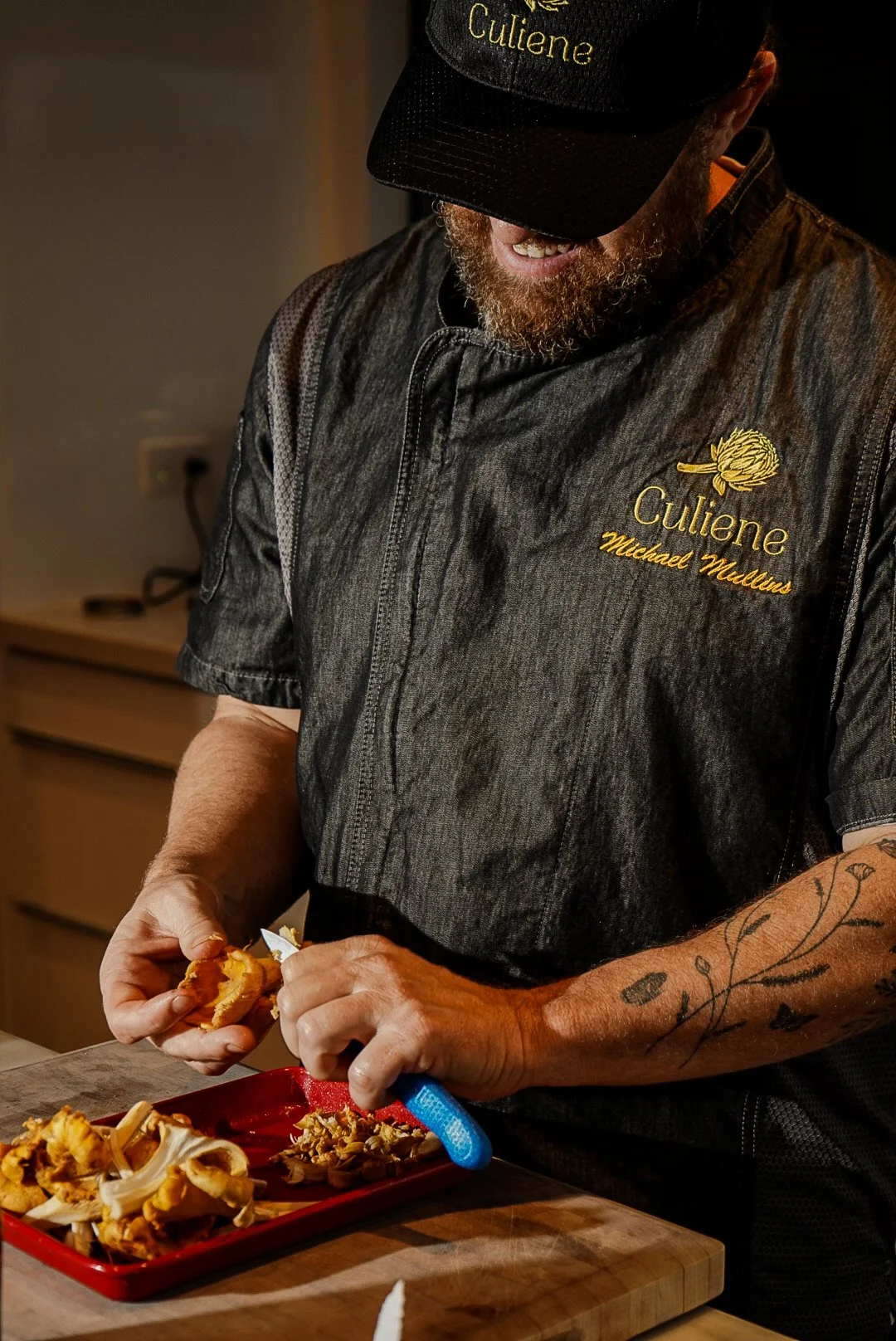 A chef wearing a black cap and apron with the name 'Culiène' and 'Michael Mullins' embroidered on it. He is peeling a vegetable or fruit over a red tray filled with cooked mushrooms and other food items in a kitchen.