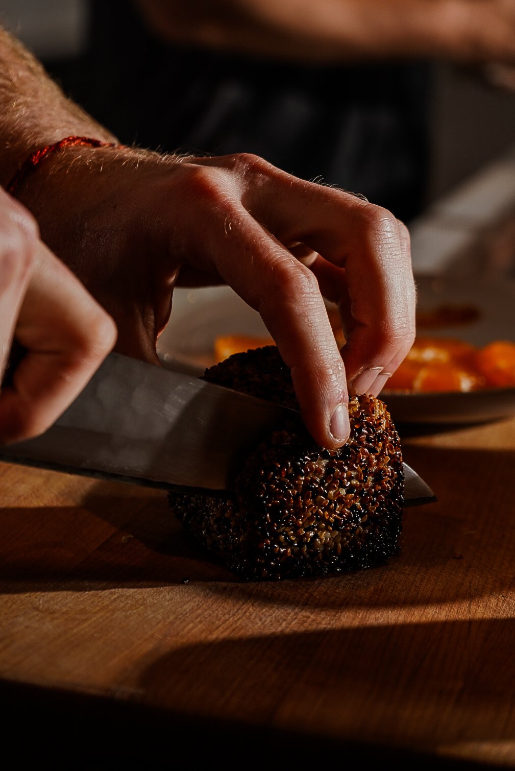 A person slicing a sesame-crusted breaded ball on a wooden cutting board with a large knife.