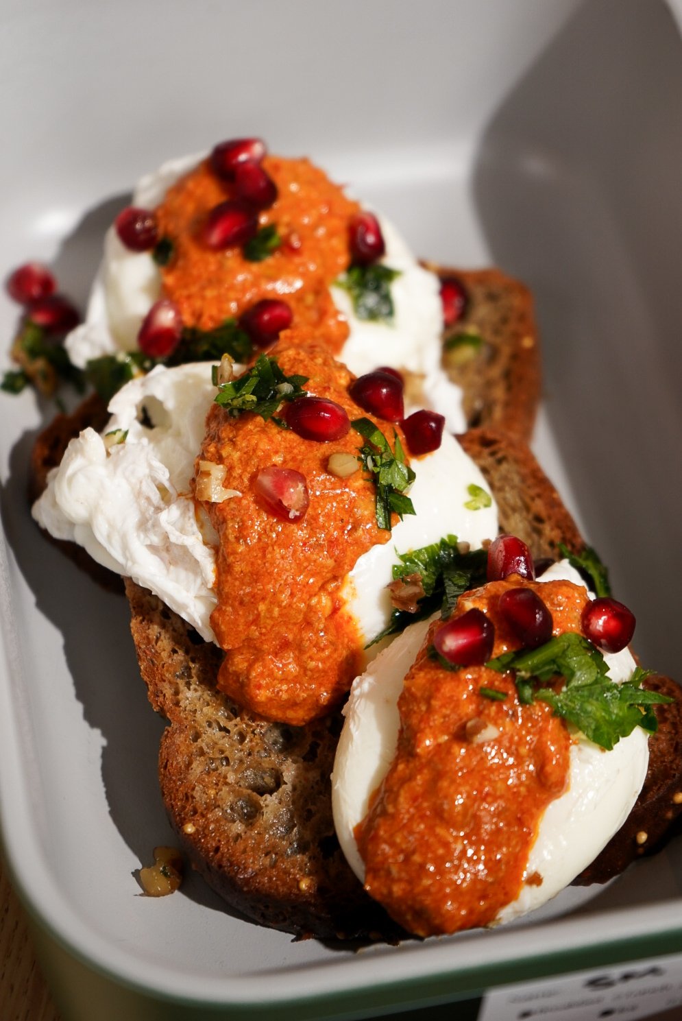 Open-faced sandwich with bread, poached eggs, tomato sauce, chopped parsley, and pomegranate seeds.