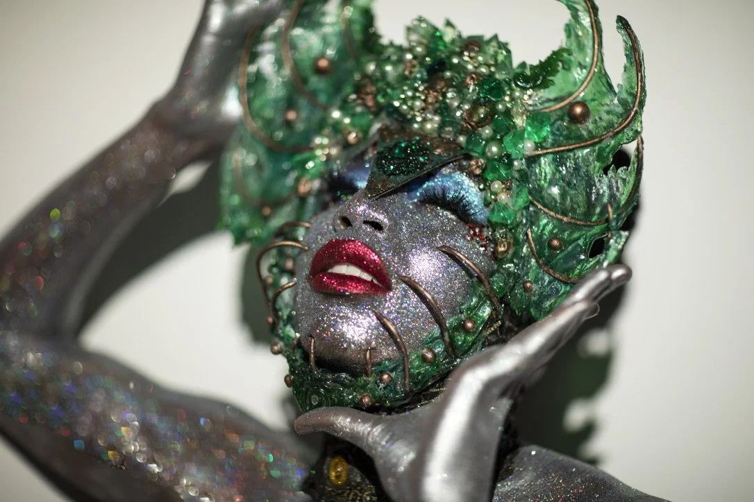 An artistic sculpture of a woman with glittery makeup, blue eyeshadow, red lipstick, and closed eyes, surrounded by green glass and copper wire, with her hands framing her face.