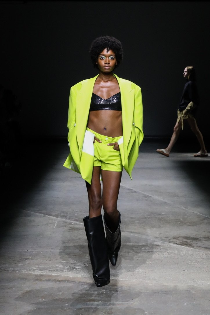 A female model walking on a runway wearing a black sequin crop top, bright yellow blazer and shorts, and black knee-high boots, with another model walking in the background.