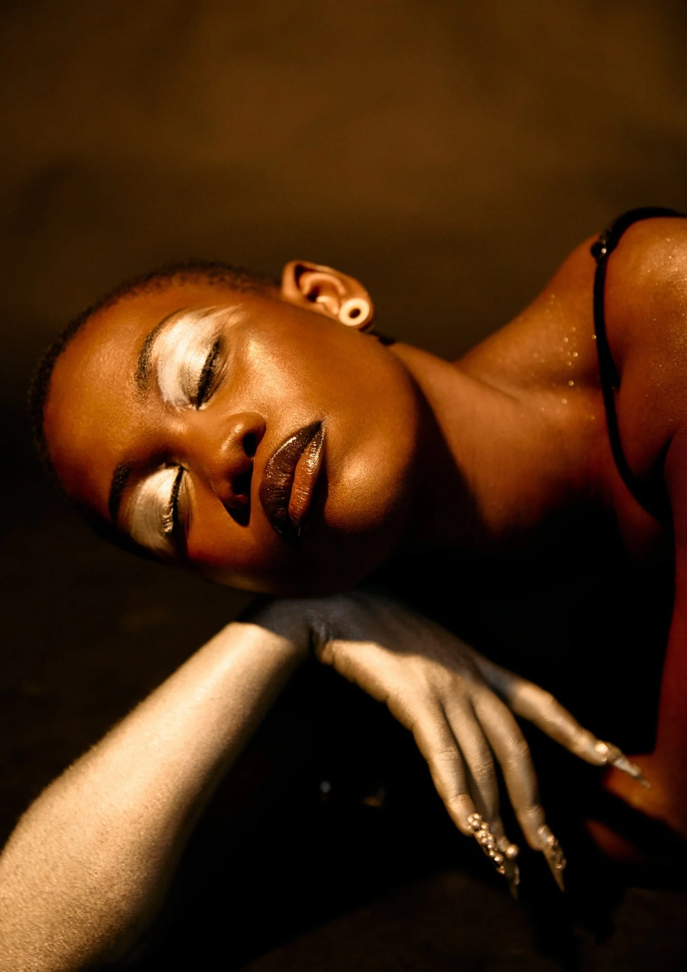 A woman with dark skin and short hair, lying down with eyes closed, wearing makeup, jewelry, and a black top, against a dark background.