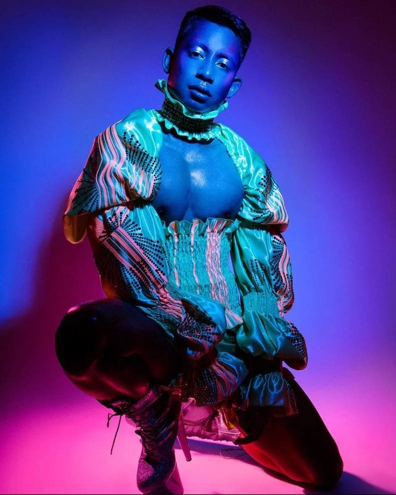 A person with short hair poses against a gradient background of purple and pink lighting. They are wearing a colorful, patterned, oversized jacket and matching pants, with one knee bent and head tilted slightly.