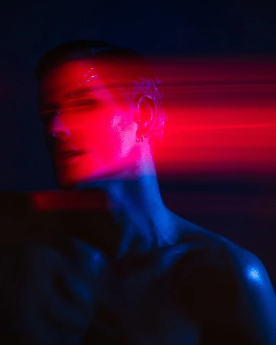A person with short hair and earrings is illuminated by pink and blue lights, with streaks of red and pink light crossing their face and background, creating a futuristic, artistic portrait.