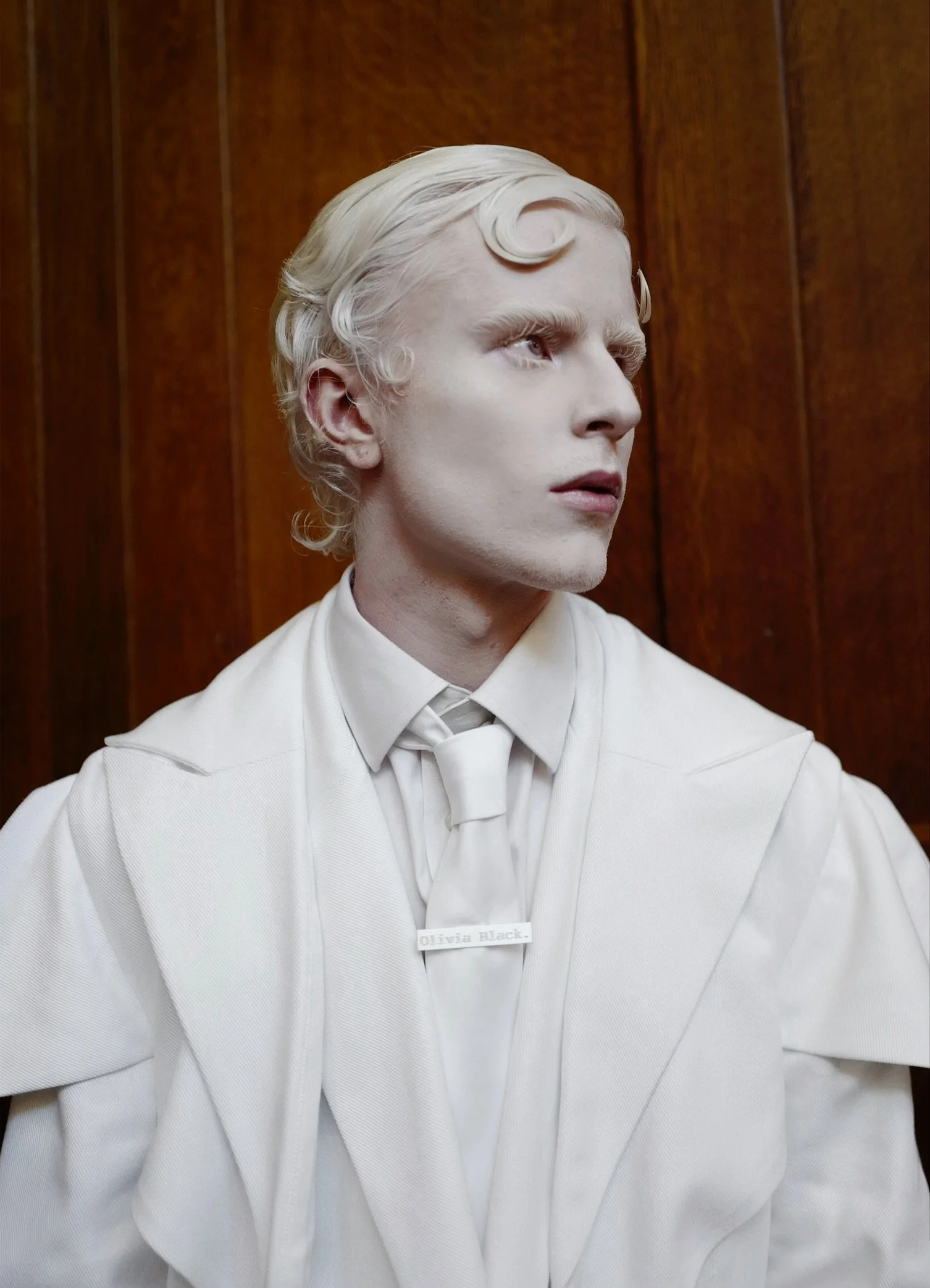 A person in a cream-colored suit with a name tag that reads 'Olivia Black' is looking to the right against a wooden panel background.