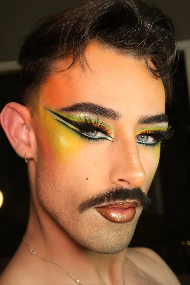 A person with dramatic makeup featuring bold eyeshadow in yellow and green, black liner, long lashes, metallic lips, and a defined mustache.