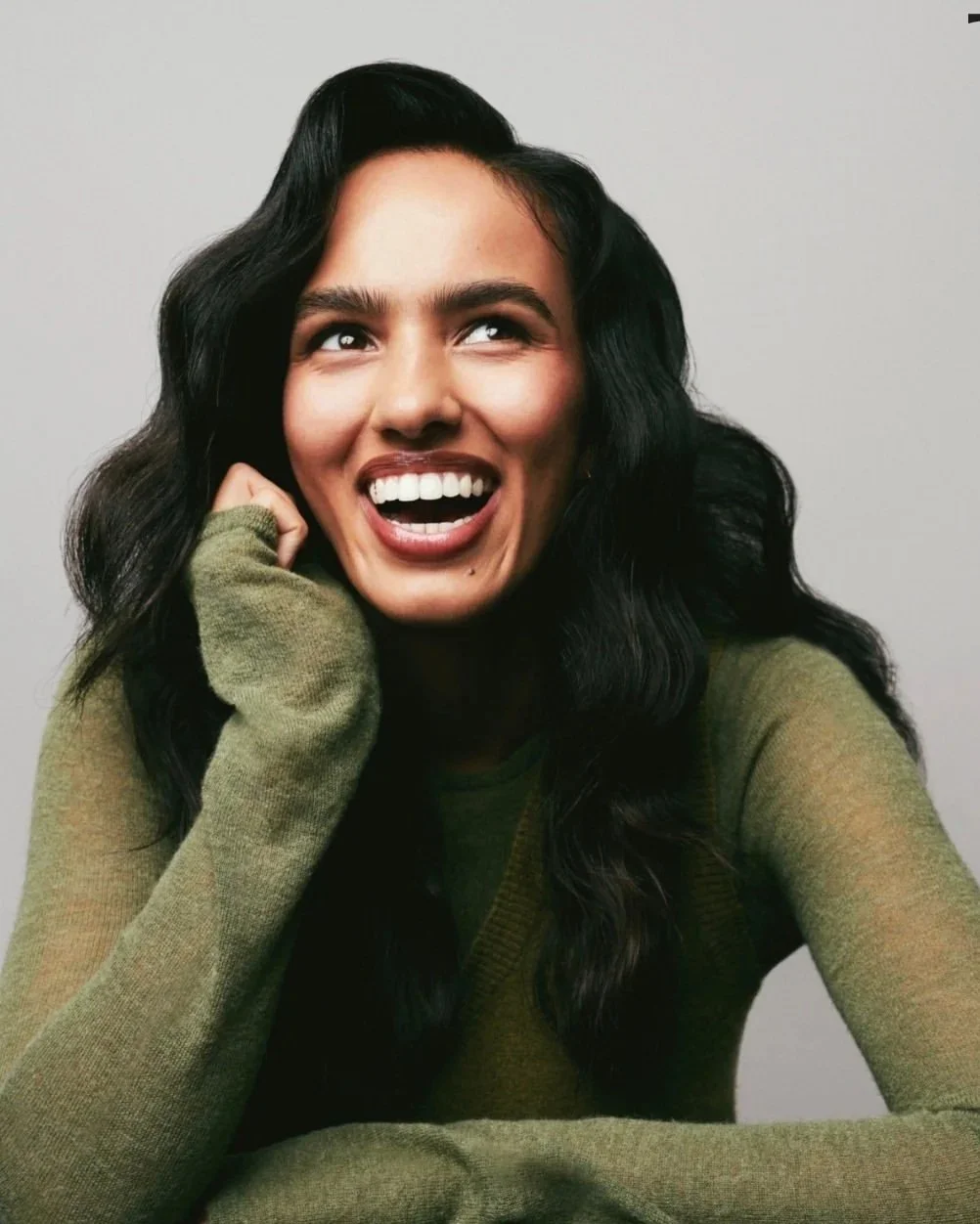 A woman with long black wavy hair smiling and looking up, wearing an olive green sweater, posing with her hand near her face against a light gray background.