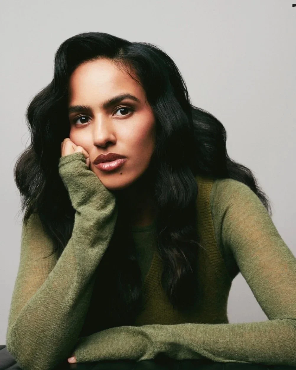 A woman with long black wavy hair and olive green sweater resting her face on her hand, looking directly at the camera against a plain background.