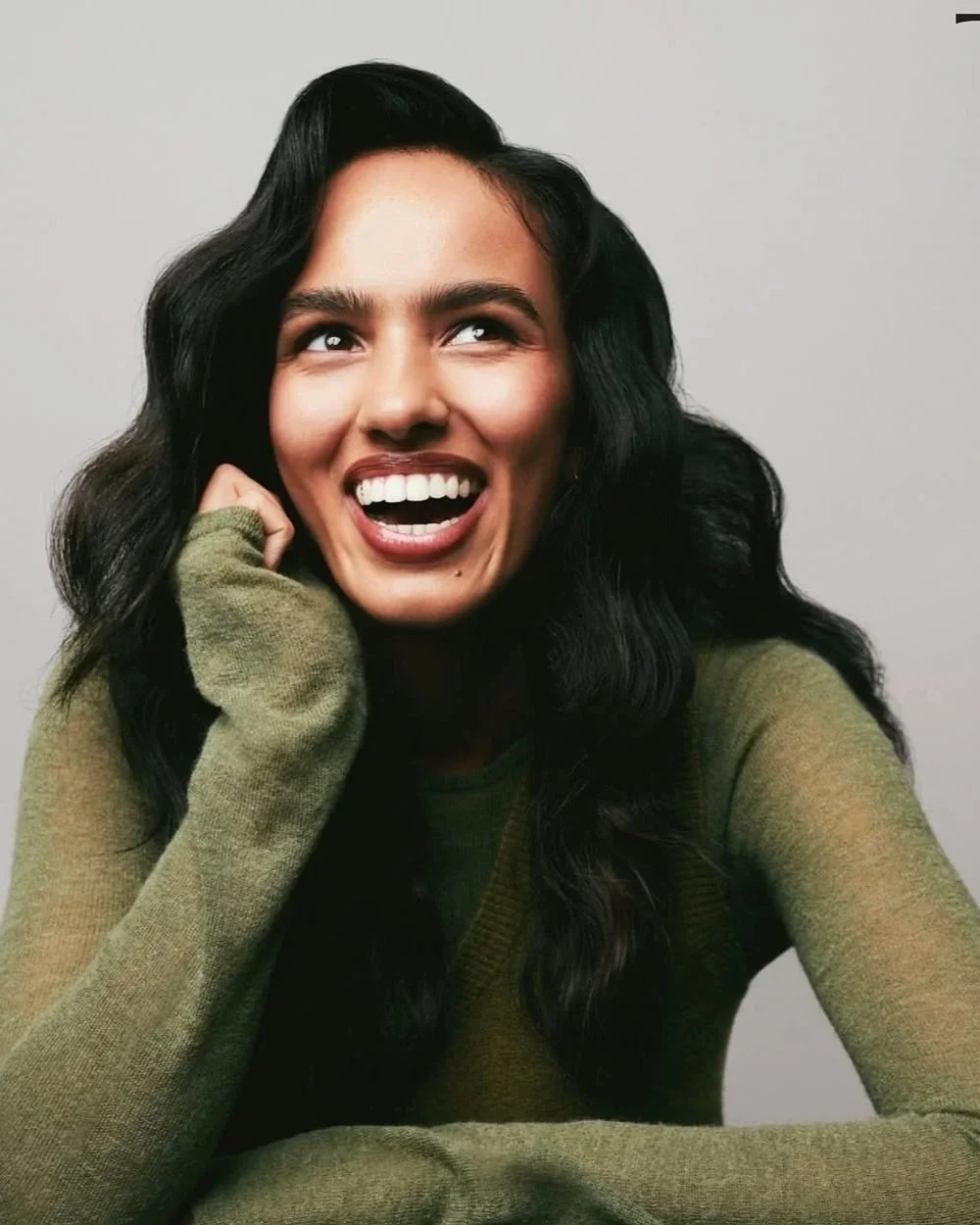A woman with long black curly hair smiling and looking up, wearing a green long-sleeve shirt, with one hand resting near her face against a plain gray background.