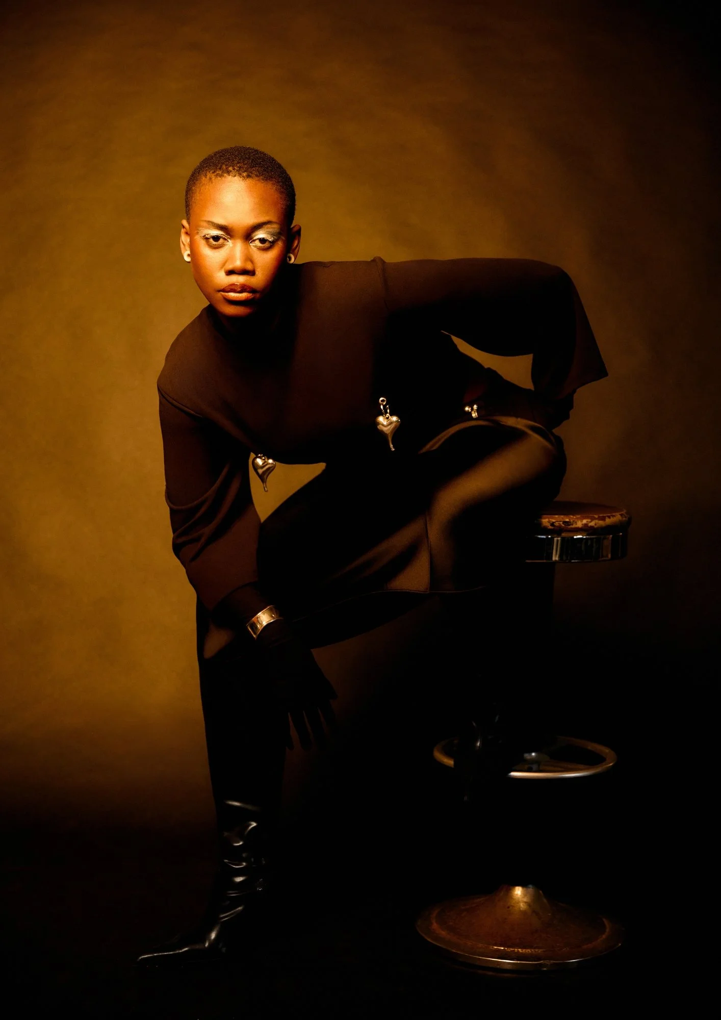 A woman with short hair and earrings is leaning forward, wearing a black top with metallic heart-shaped embellishments. She is positioning her left leg on a high stool, with her right leg extended downward. She is wearing shiny black boots. The backg