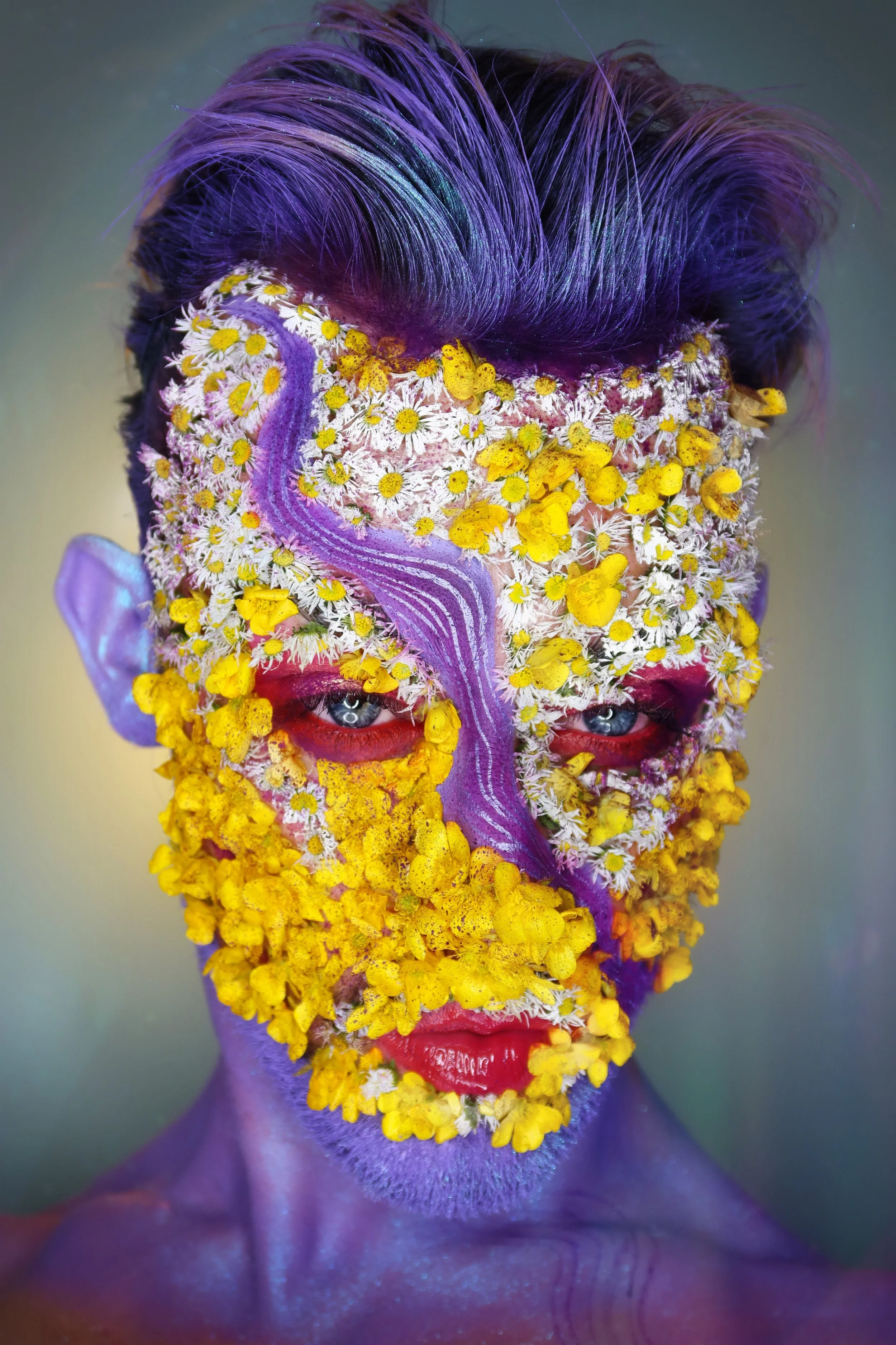 A portrait of a person with face and body painted purple, adorned with yellow and white flowers and purple streaks, with eyes looking directly forward.