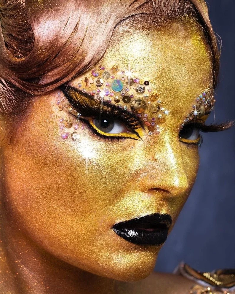 Close-up of a person's face painted and adorned with gold makeup and rhinestones, with dramatic eye makeup including black eyeliner and long eyelashes.