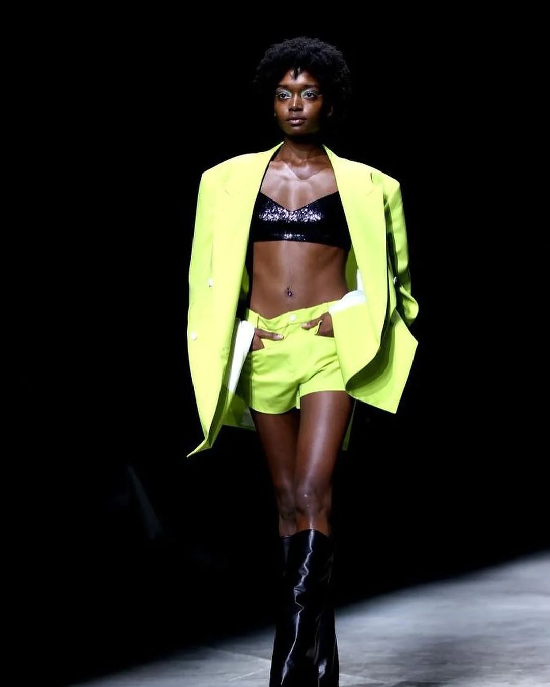 A fashion model walking on the runway in a black and neon yellow outfit, with a black sequined crop top, matching neon yellow shorts, a bright yellow blazer, and black knee-high boots, against a dark background.
