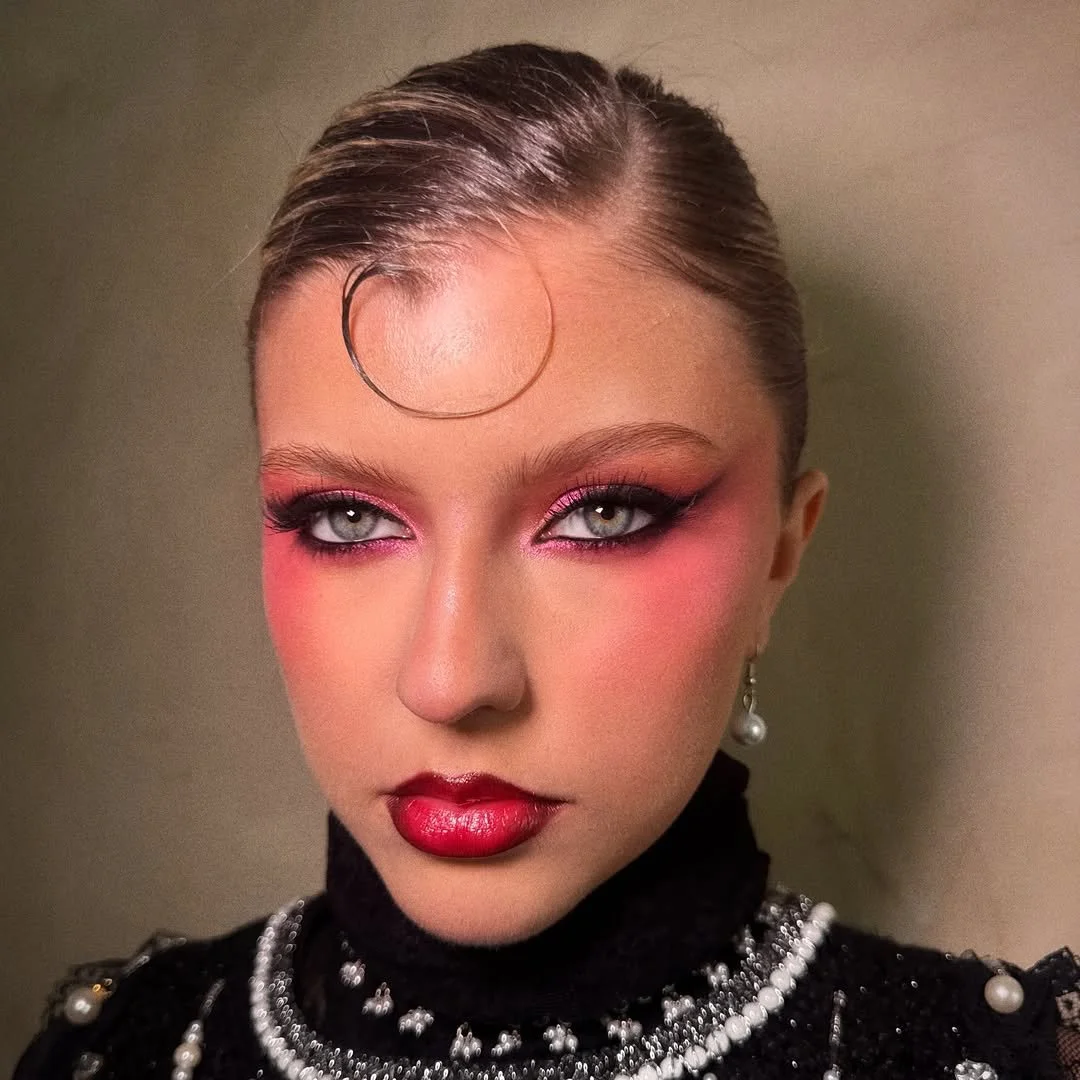 A woman with slicked-back hair, bold pink and red makeup, and a black embellished top with pearl accessories.
