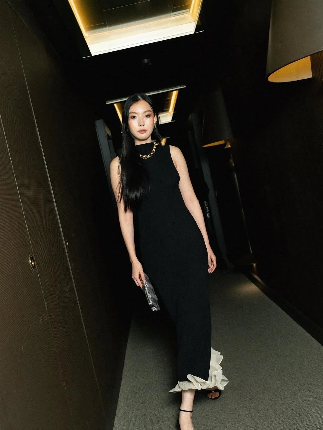 Woman in a black sleeveless dress with a beige ruffle at the bottom, standing in a dark hallway with modern lighting.