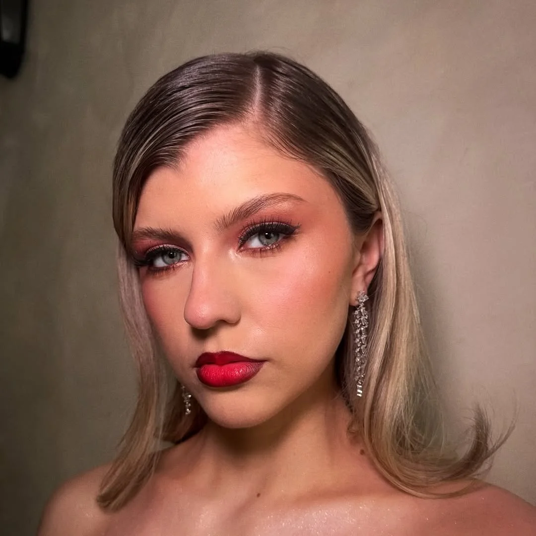 A close-up portrait of a woman with blonde hair, blue eyes, and red lipstick, wearing chandelier earrings.