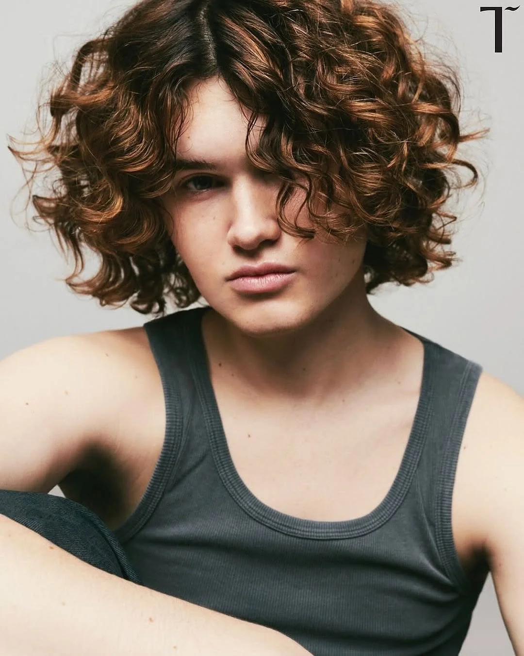 A woman with curly reddish-brown hair partially covering her face, wearing a dark sleeveless top, staring with a serious expression against a plain background.