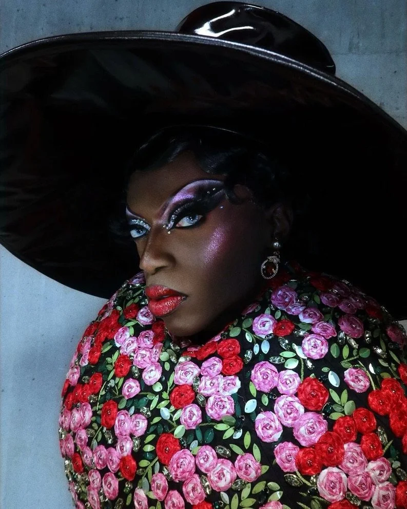 A woman wearing a large black hat, colorful floral embroidered jacket, and dramatic makeup with sparkly eyeshadows and red lipstick.