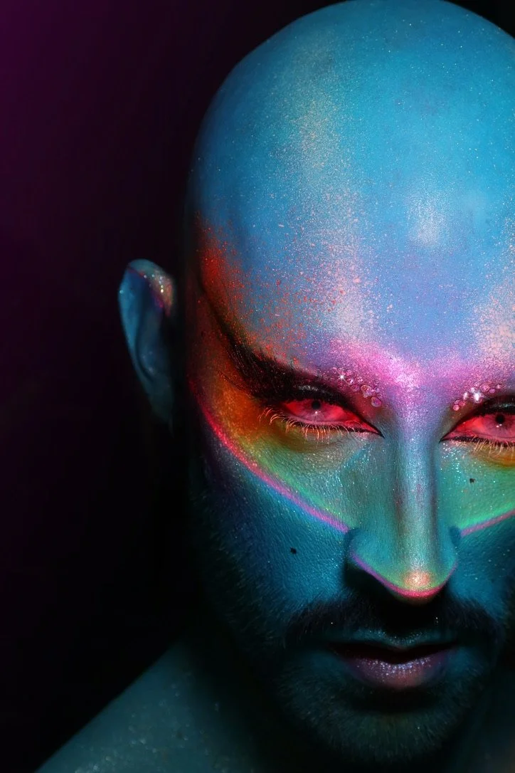 A bald person with glowing, colorful makeup and paint on their face, featuring neon shades of pink, purple, blue, green, and orange against a dark background.