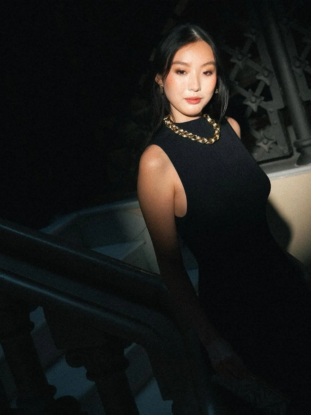 A woman with long black hair wearing a sleeveless black dress with gold jewelry, sitting inside a vehicle.