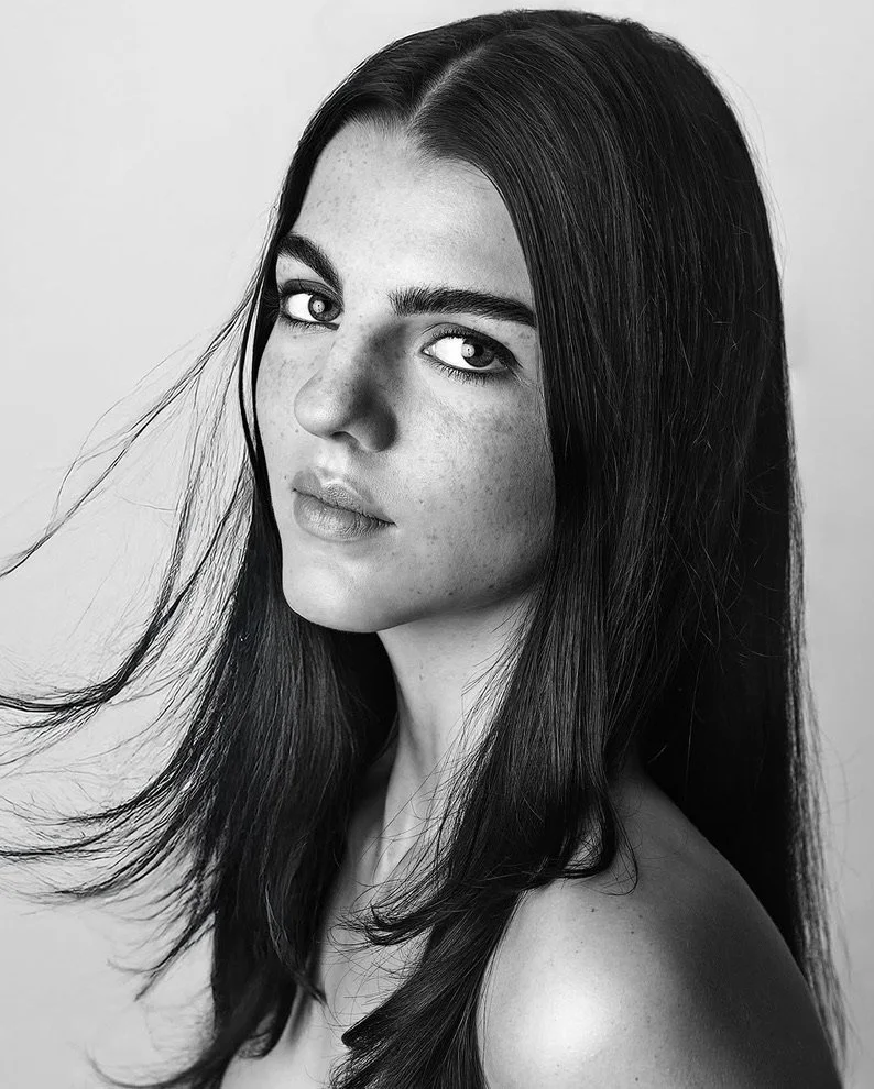 Black and white portrait of a woman with long dark hair, light skin with freckles, gazing directly at the camera with a neutral expression.