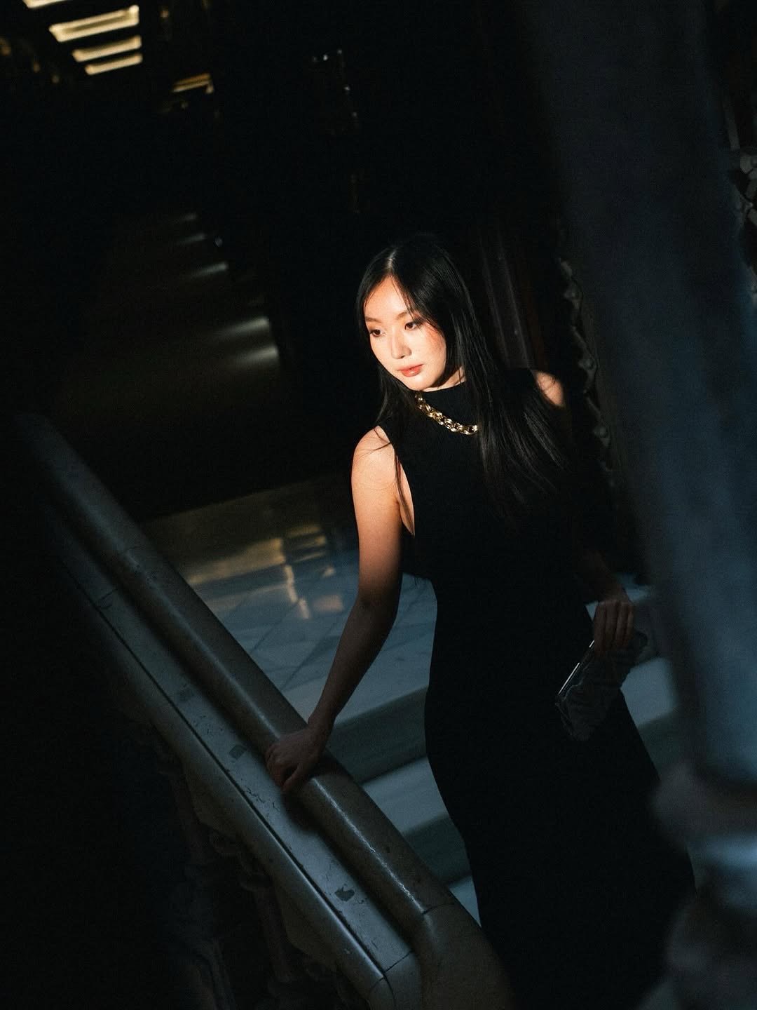 A woman with long black hair in a black sleeveless dress with a gold chain necklace, standing on an escalator in a dark indoor setting, holding onto the escalator handrail.