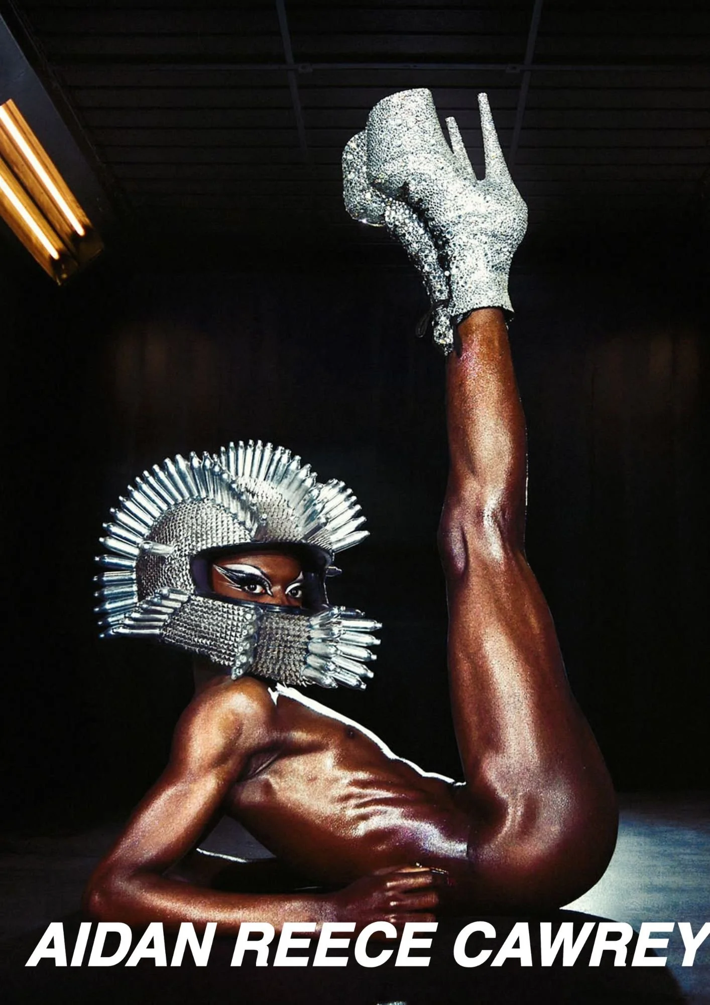 A person with dark skin, wearing a silver metallic headdress and matching face mask, poses with a raised arm and a finger pointing upward. The person's body is body painted in a shiny, metallic color, and the background is dark with the text "AIDAN R
