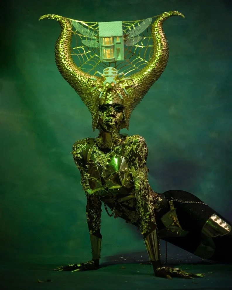 A humanoid figure with a female appearance, covered in reflective and metallic materials, kneeling on the ground in front of a green background. The figure has an intricate headpiece resembling a spider web with a house and a spider, blending organic