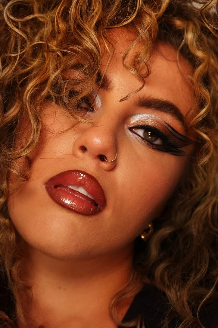 A woman with curly blonde hair, metallic eye makeup, glossy lips, and a nose ring.