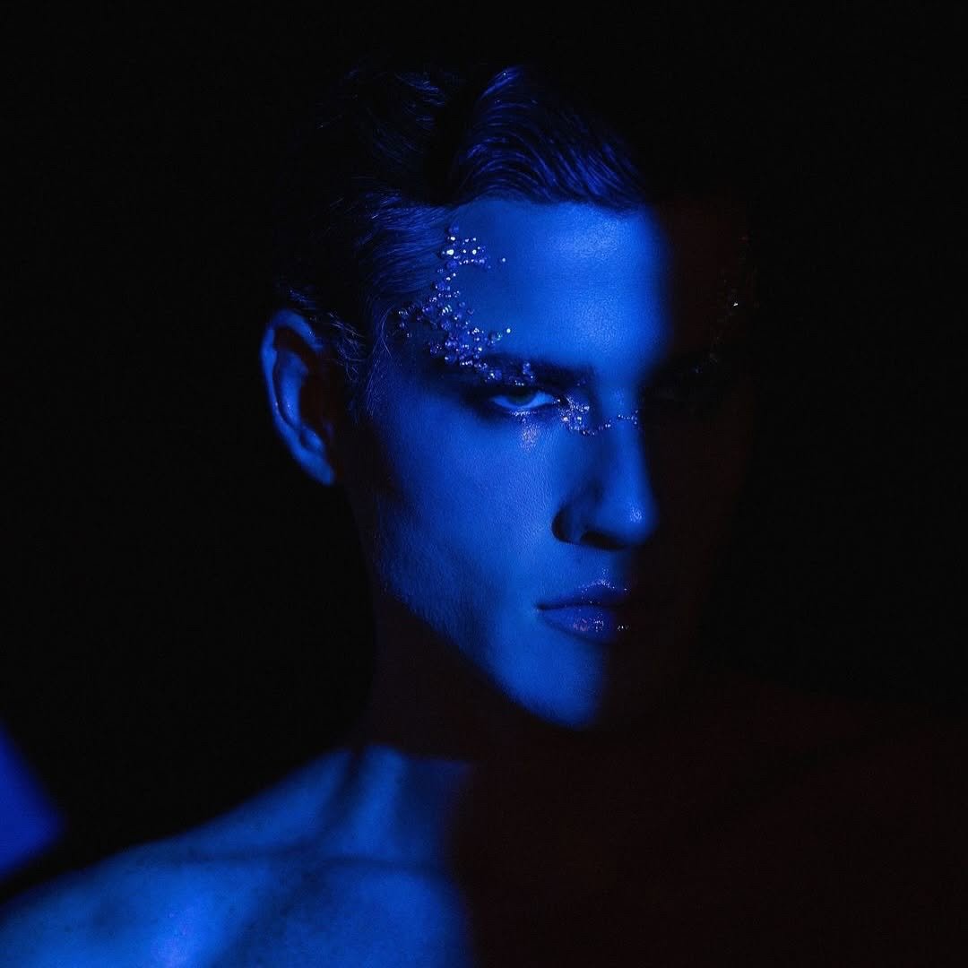 A person with short hair and metallic makeup, illuminated with blue lighting, looking at the camera in a dark setting.