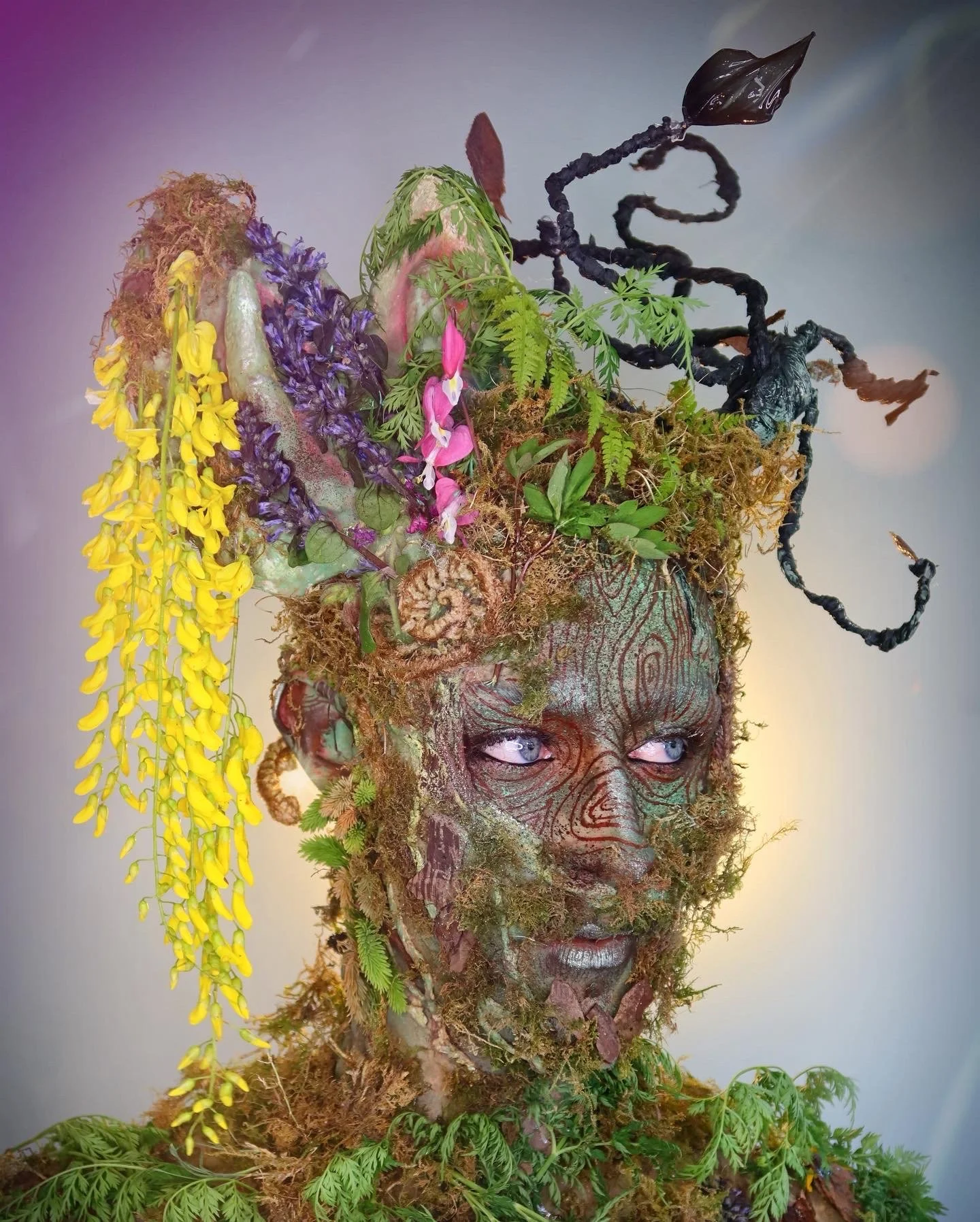 A detailed artistic sculpture of a human face made from natural materials such as moss, leaves, and flowers, with prominent blue eyes and vibrant plant decorations on the head resembling hair.