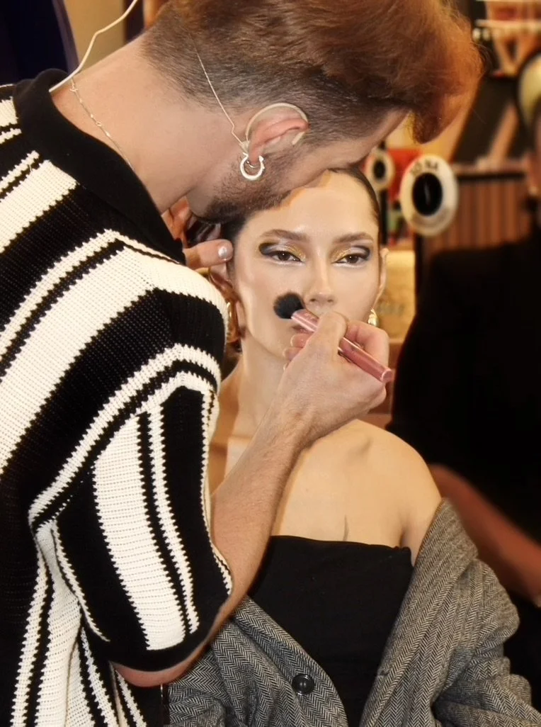 A makeup artist is applying makeup on a woman with short dark hair and gold eyeshadow, wearing a strapless black top and a gray blazer, in a brightly lit room.