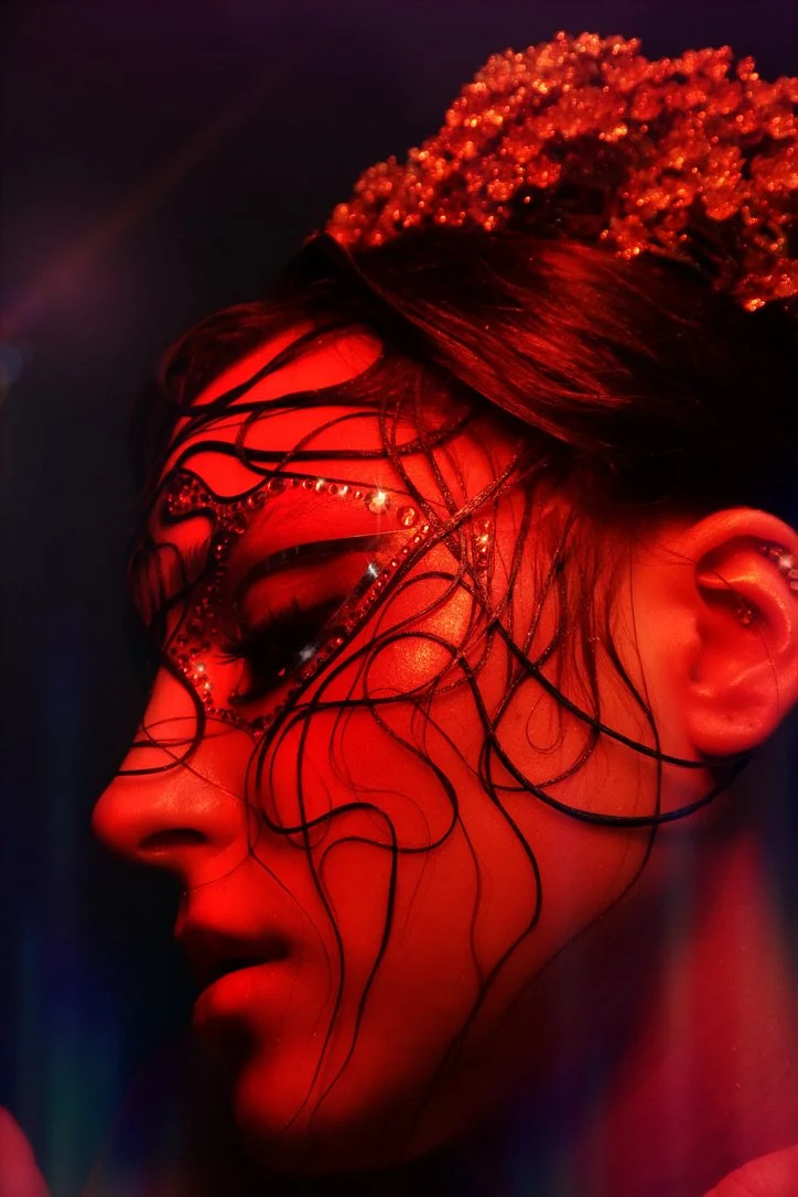 A woman with wet hair, illuminated by red lighting, features elaborate crystal and black wire face decorations, a pearl headpiece, and an earring in her ear.