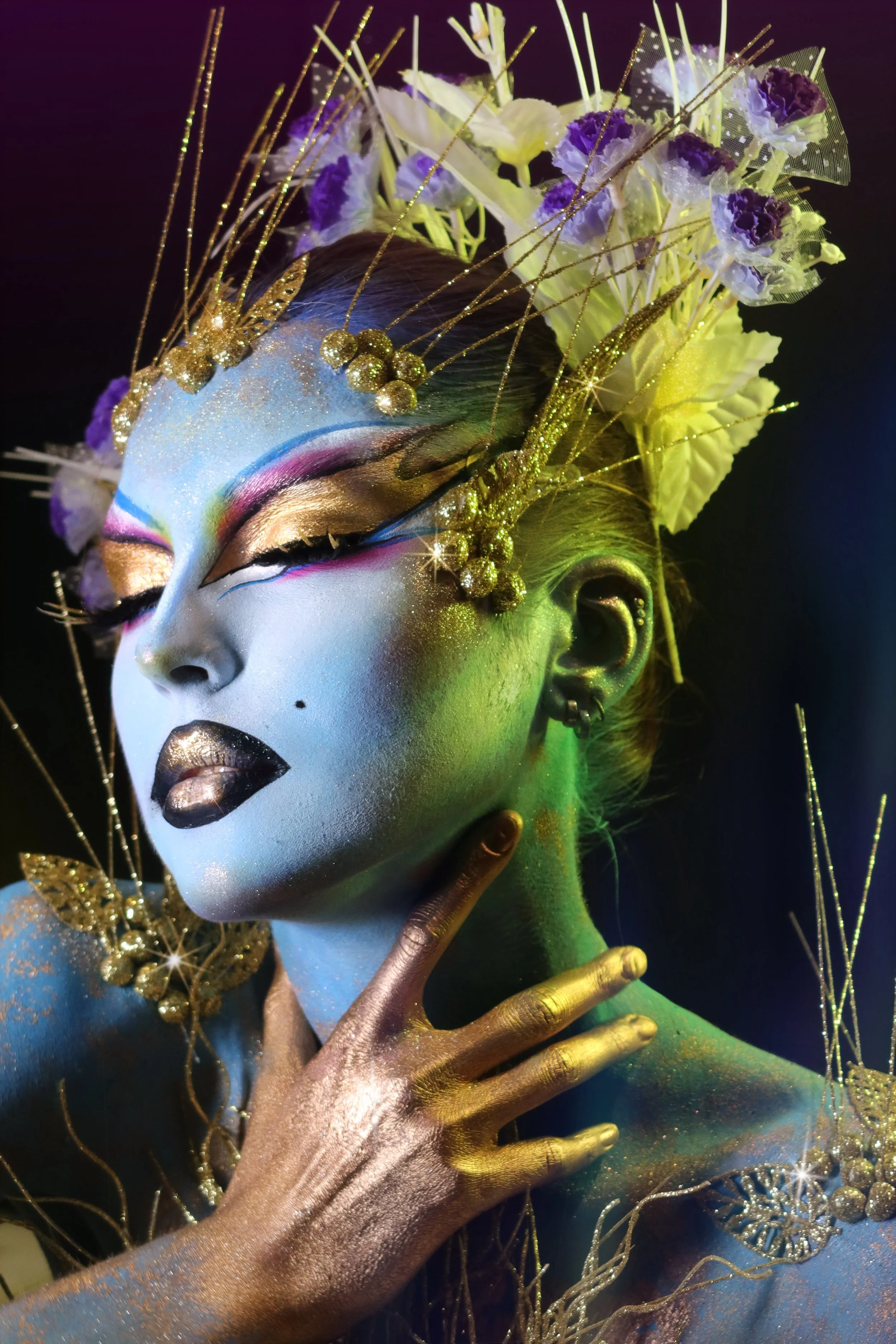A person with colorful, artistic makeup and gold-painted skin, adorned with glitter and golden jewelry, wearing a headpiece made of purple and white flowers, gold twigs, and netted fabric.