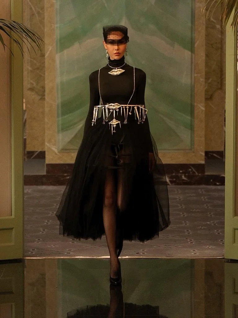 A woman in a black turtleneck dress with sheer black skirt walking forward, wearing a black veil on her head, black high heels, and elaborate silver jewelry.