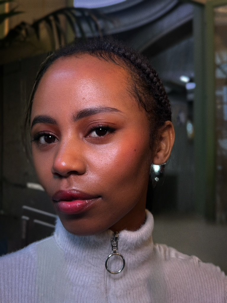 Close-up portrait of a young woman with braided hair and glowing skin, wearing a gray high-neck top with a zipper.