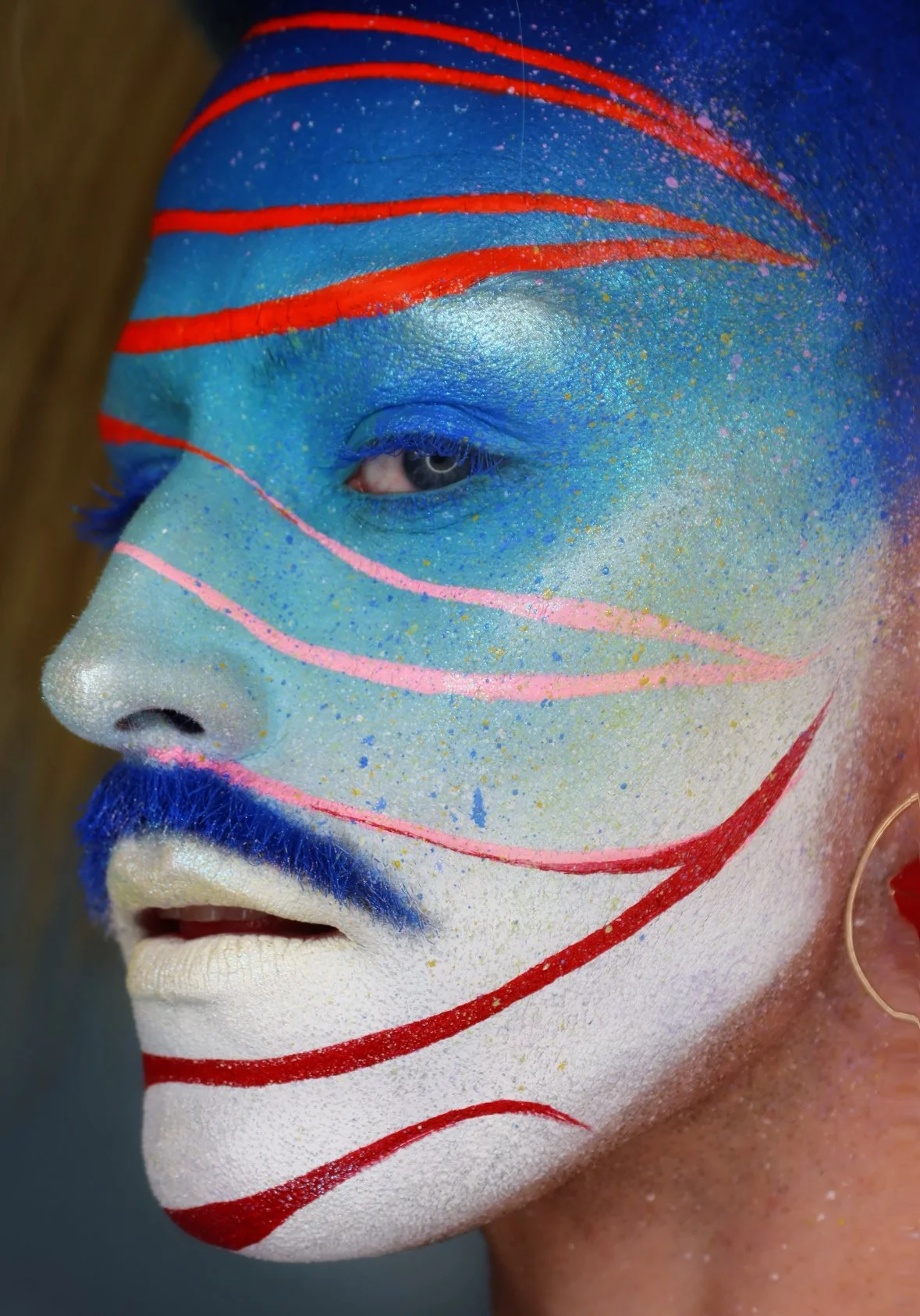 Close-up of a person's face painted in vibrant colors with red, pink, blue, and white patterns, including glitter details and a blue beard.