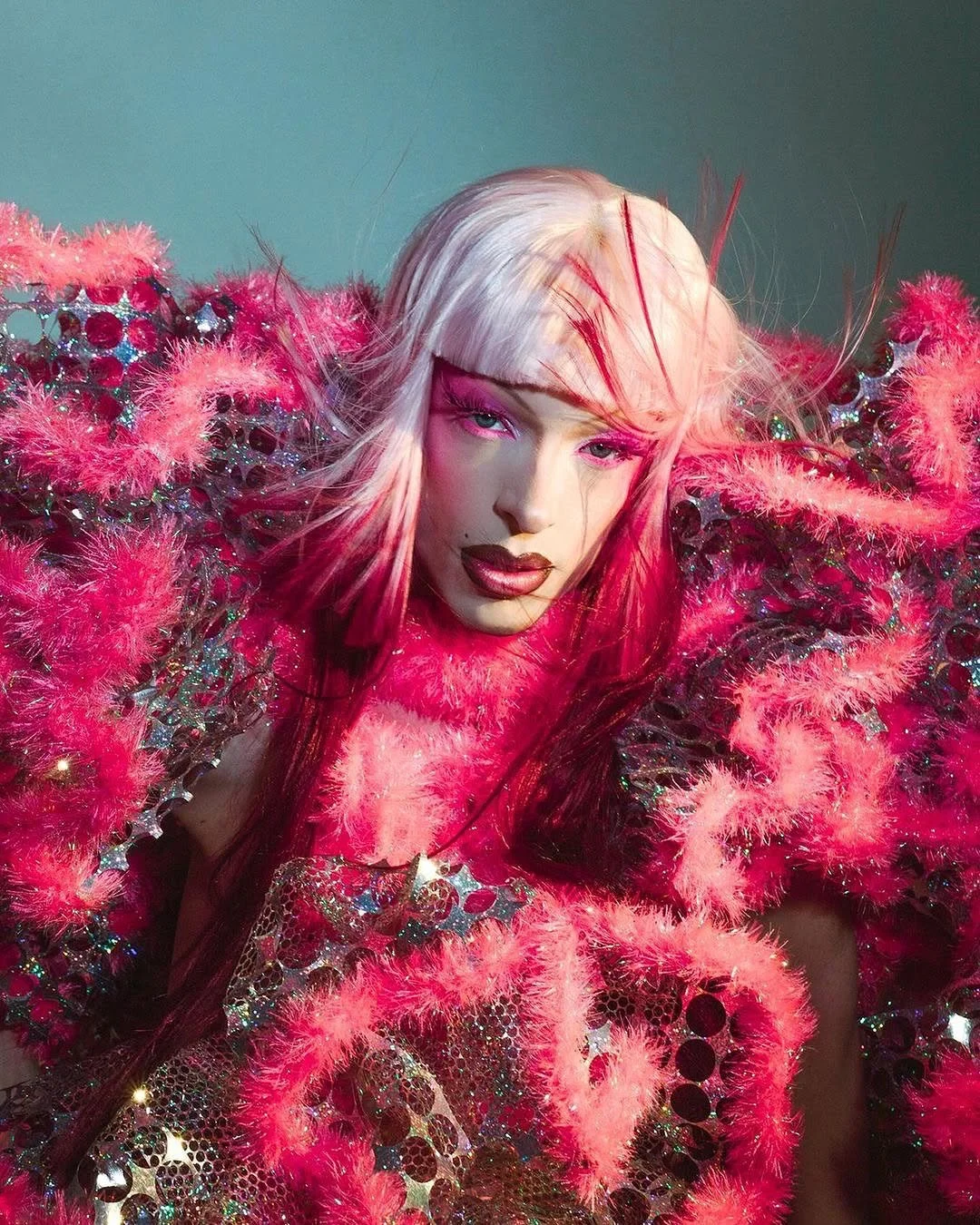 A person with platinum blonde hair, wearing an elaborate outfit made of pink feathers, sequins, and reflective materials, with vibrant pink eye makeup and lipstick.